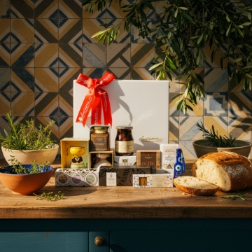 Luxury Greek Gourmet Gift Box | Premium Artisanal Foods Artisanal Greek food hamper in a rustic kitchen setting. The luxury white gift box with a red bow is on a wooden counter, surrounded by gourmet Greek products, fresh herbs, olive branches, and artisanal bread.