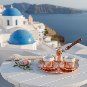 Handcrafted copper Greek coffee set on a white table overlooking the blue domes of Santorini, representing traditional Hellenic luxury and slow living.