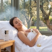 A young woman relaxing in a serene, sunlit spa environment with large windows, eyes closed, holding the Mastic Spa eye gel bottle, surrounded by nature and trees.